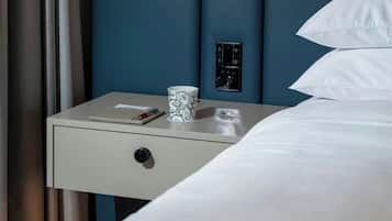 A neatly made bed with white linens, a bedside table with a lamp, a cup, and a remote control.