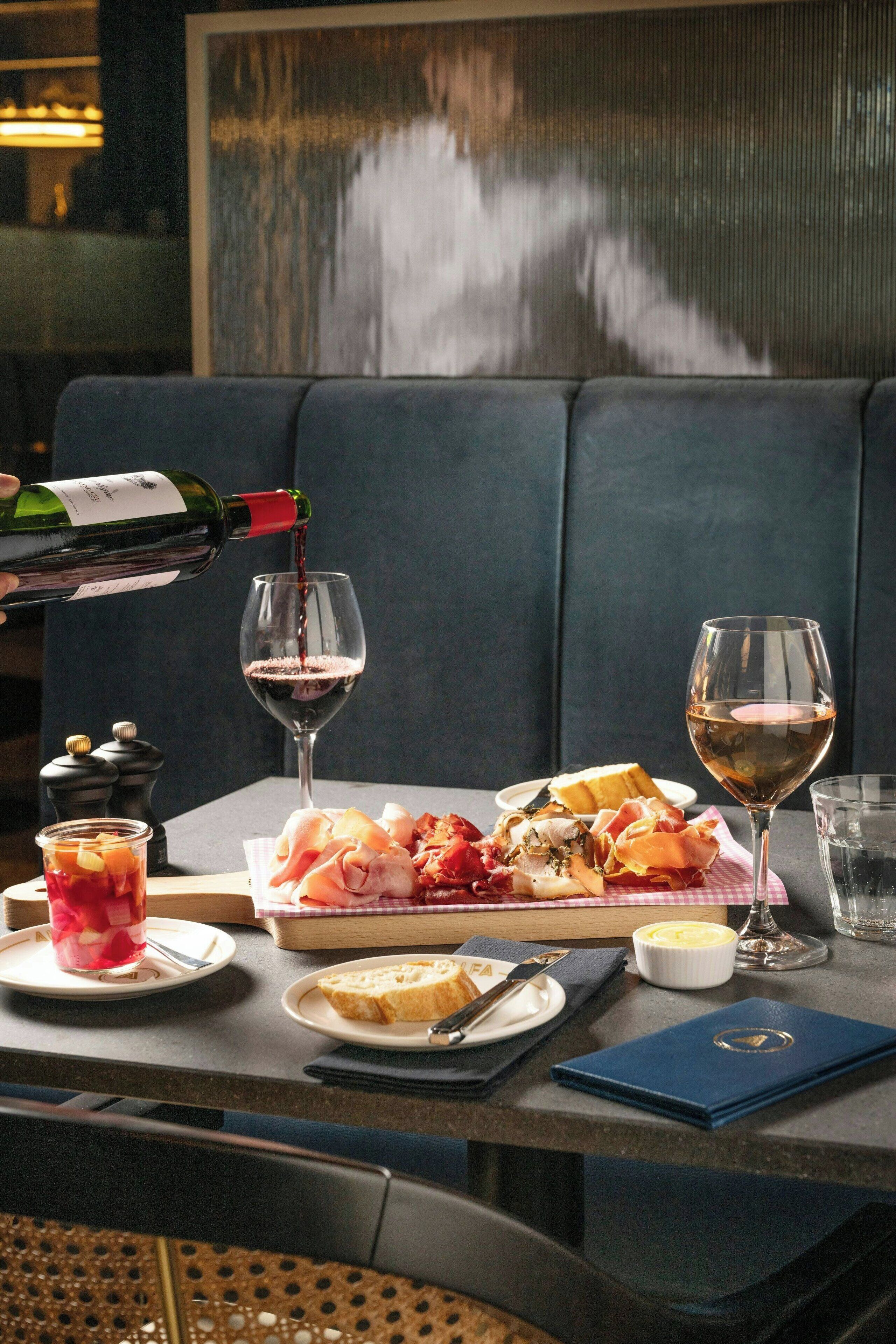 Une table à manger avec une bouteille de vin dont on verse le contenu dans un verre, une assiette de nourriture et un verre d’eau.