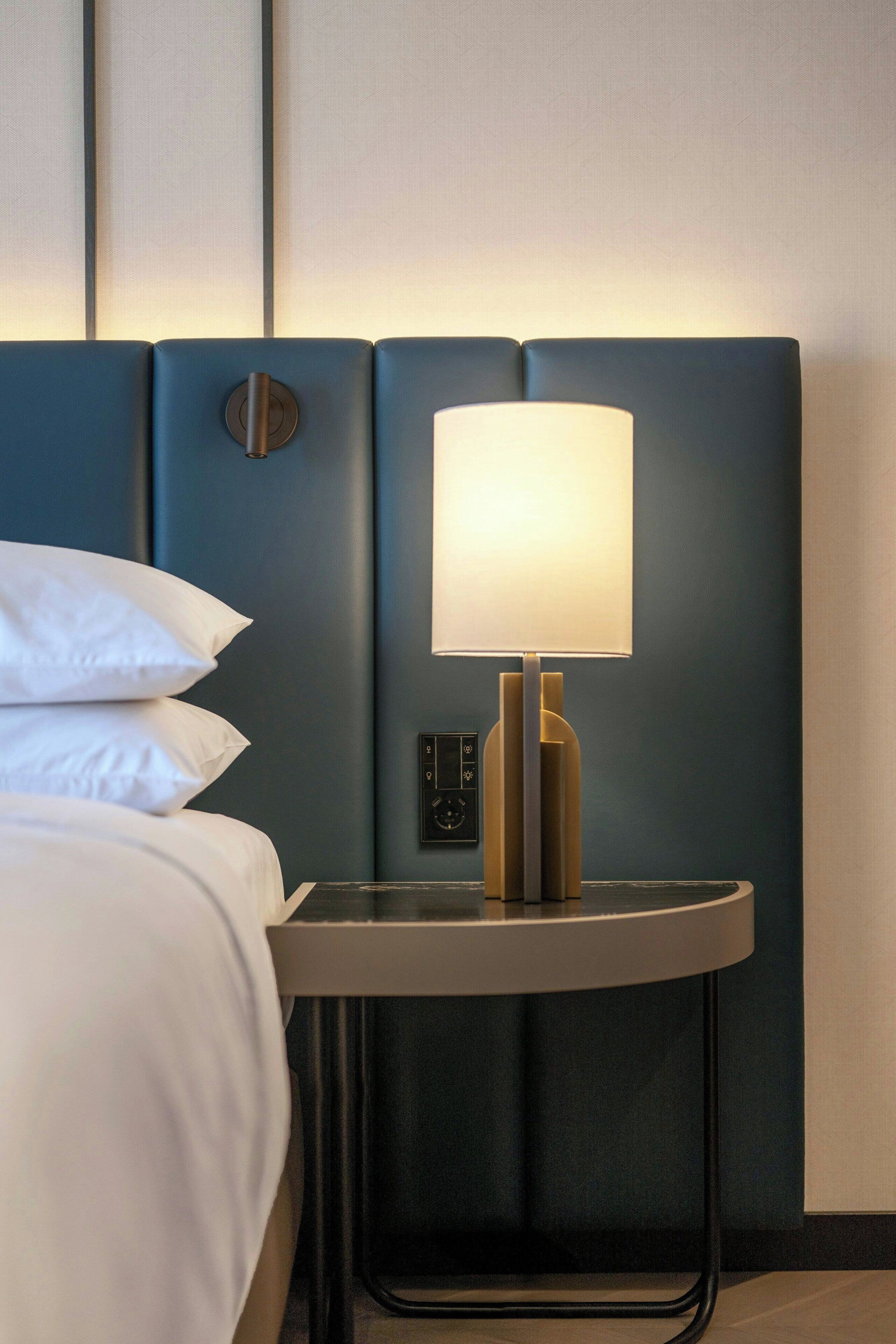 Una camera d'albergo con un letto, un comodino con una lampada e un pannello di controllo a parete.