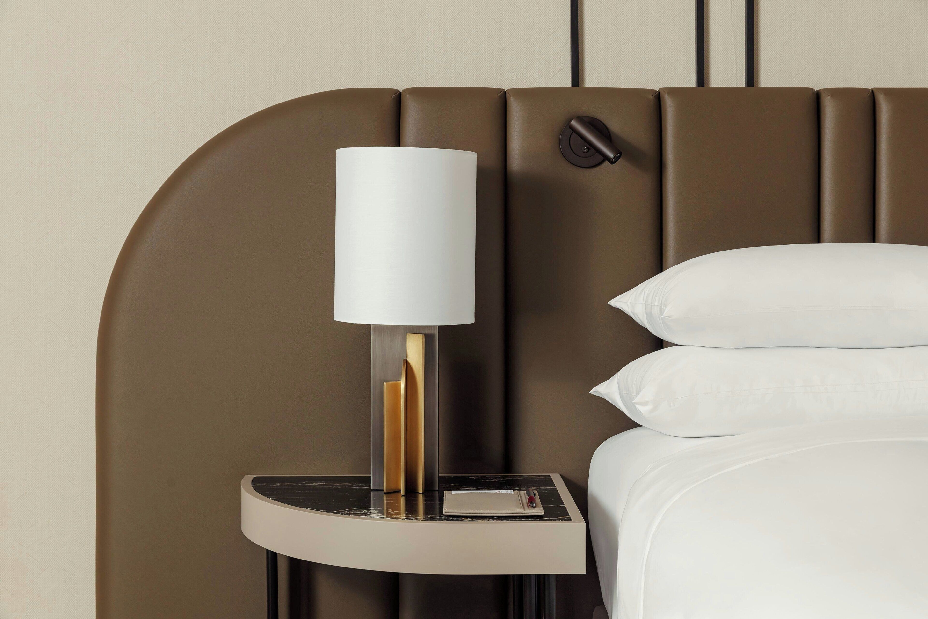 A modern hotel room with a curved headboard, a bedside table with a lamp, and neatly made beds.