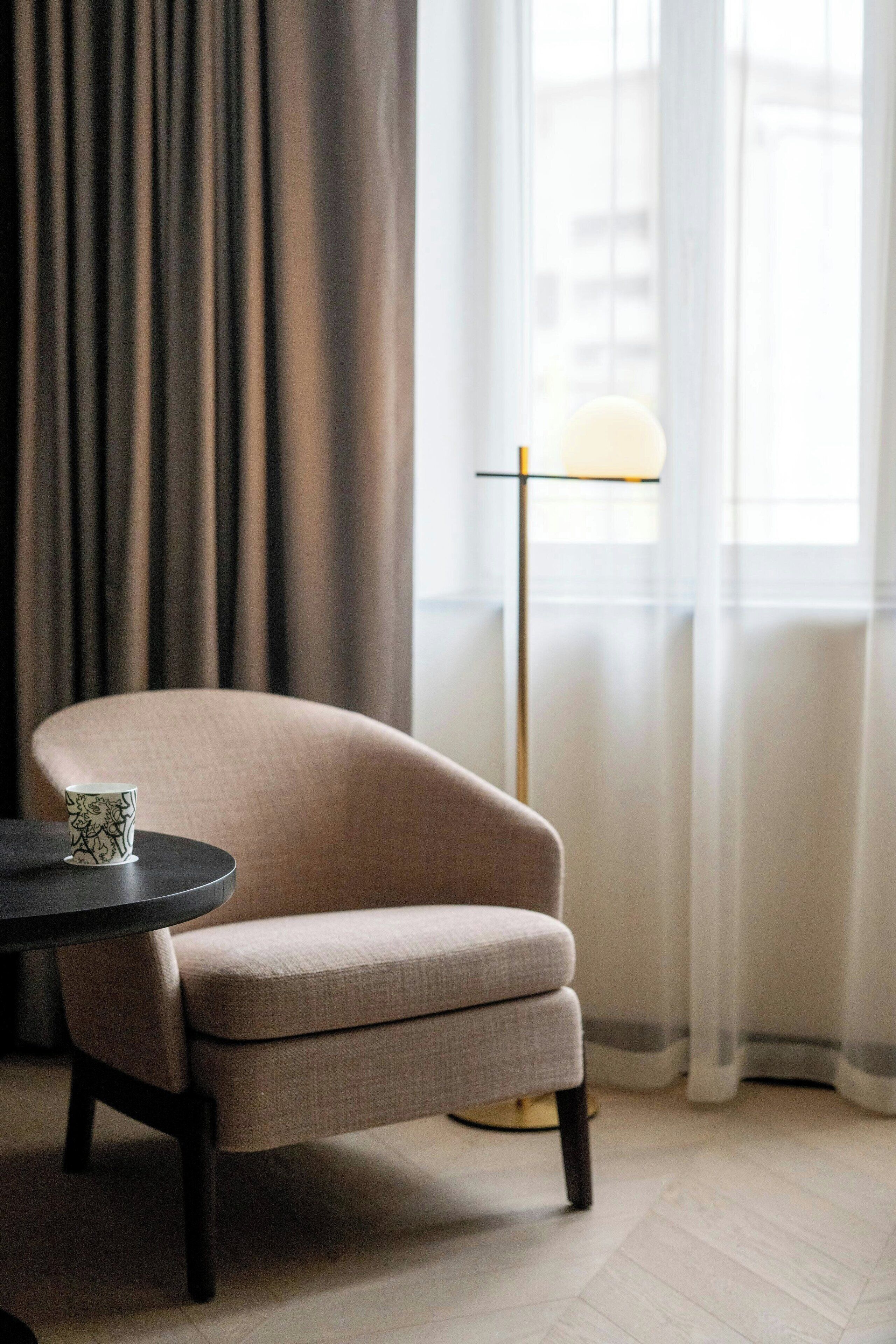 Una habitación con un sillón color crema, una mesa redonda, una lámpara de pie y vistas a través de cortinas translúcidas.