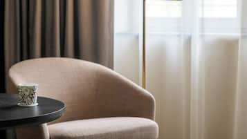 A room with a beige armchair, a round table, a floor lamp, and a view through sheer curtains.