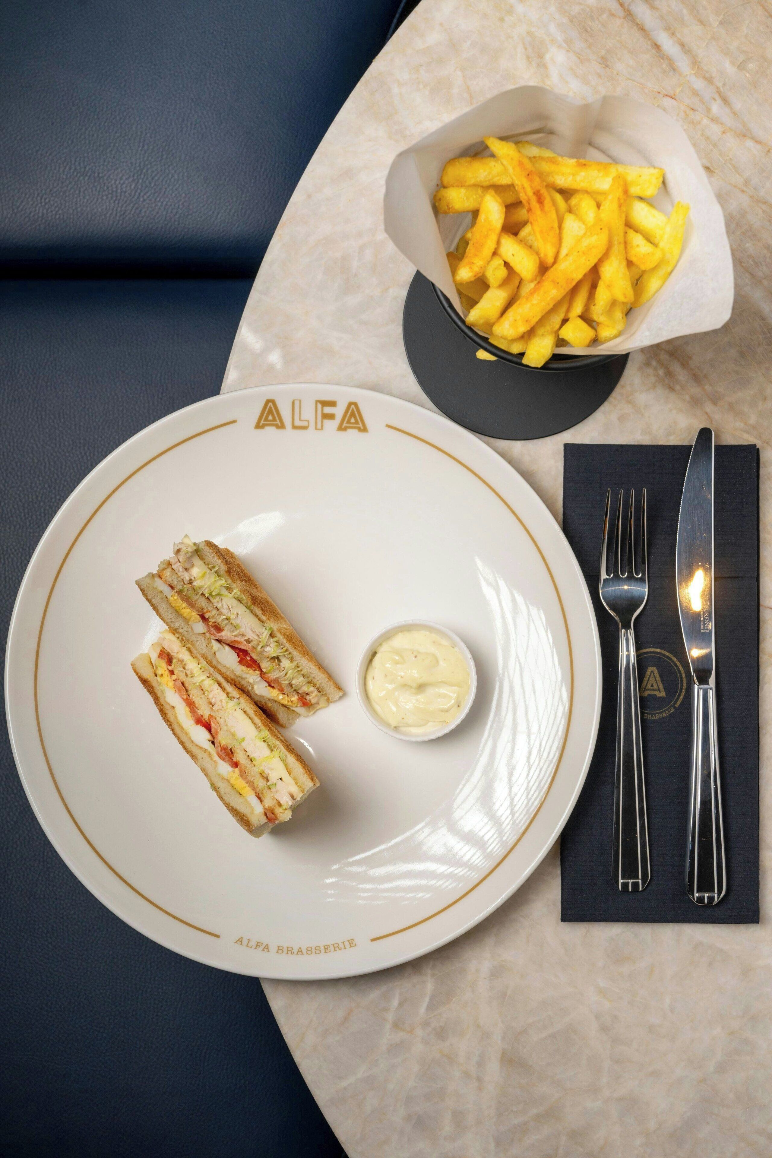Un plato con dos sándwiches y un recipiente pequeño de salsa, una cesta de papas fritas y cubiertos sobre una mesa.