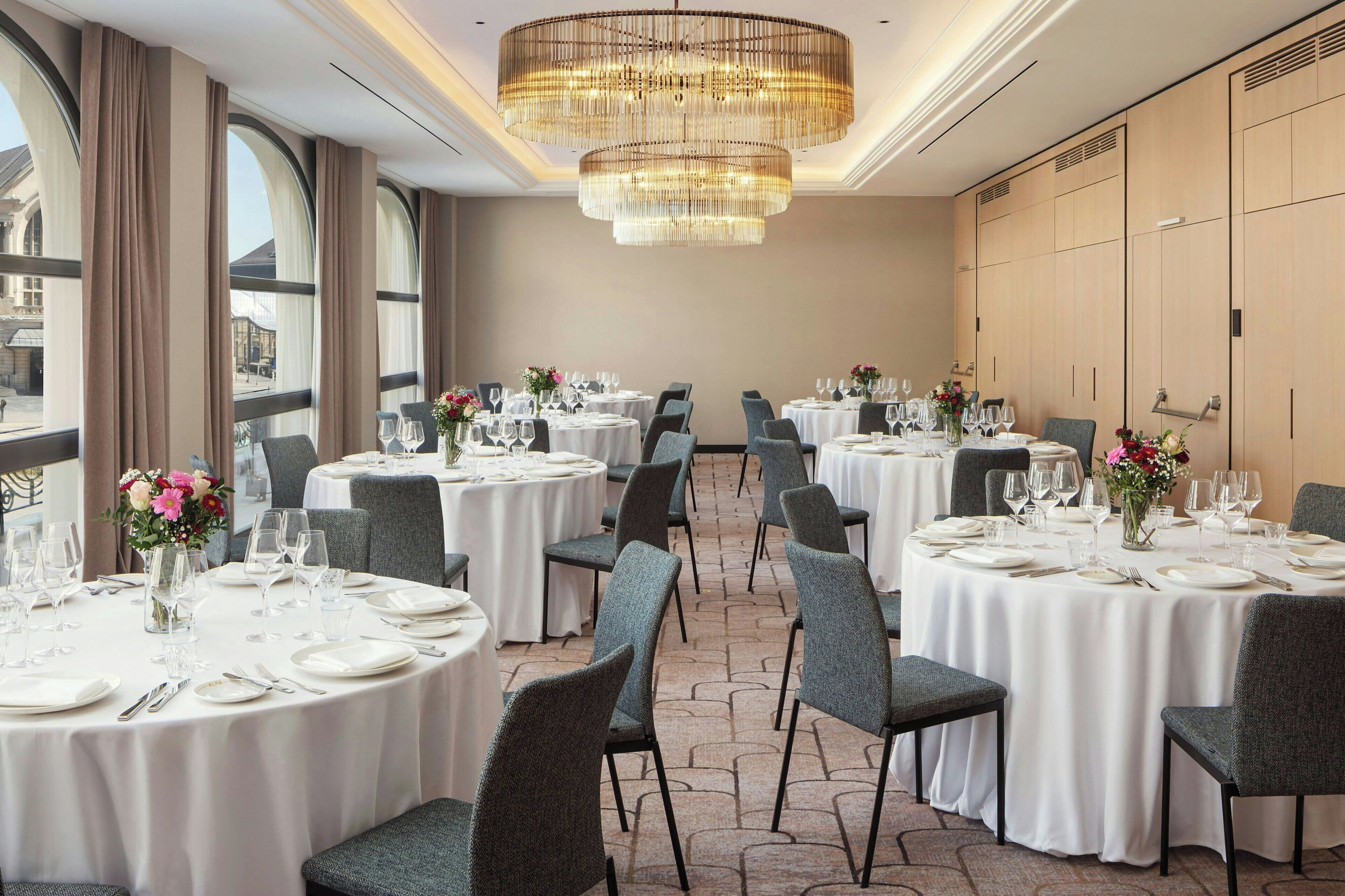Un comedor de hotel con mesas redondas dispuestas para comer, ventanas amplias y un candelabro.