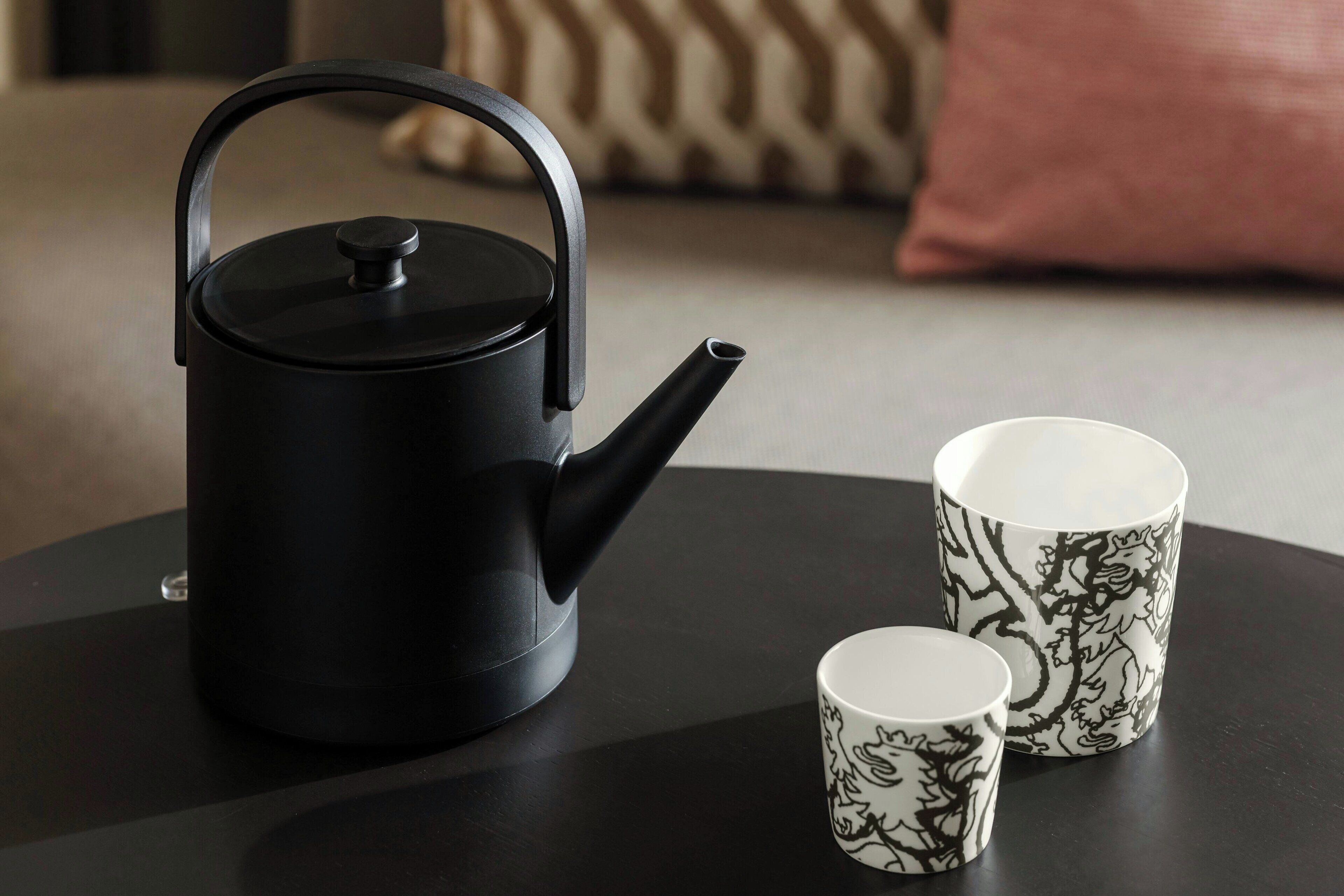 A black kettle with a lid and handle on a round table, accompanied by two white mugs with black dragon designs.