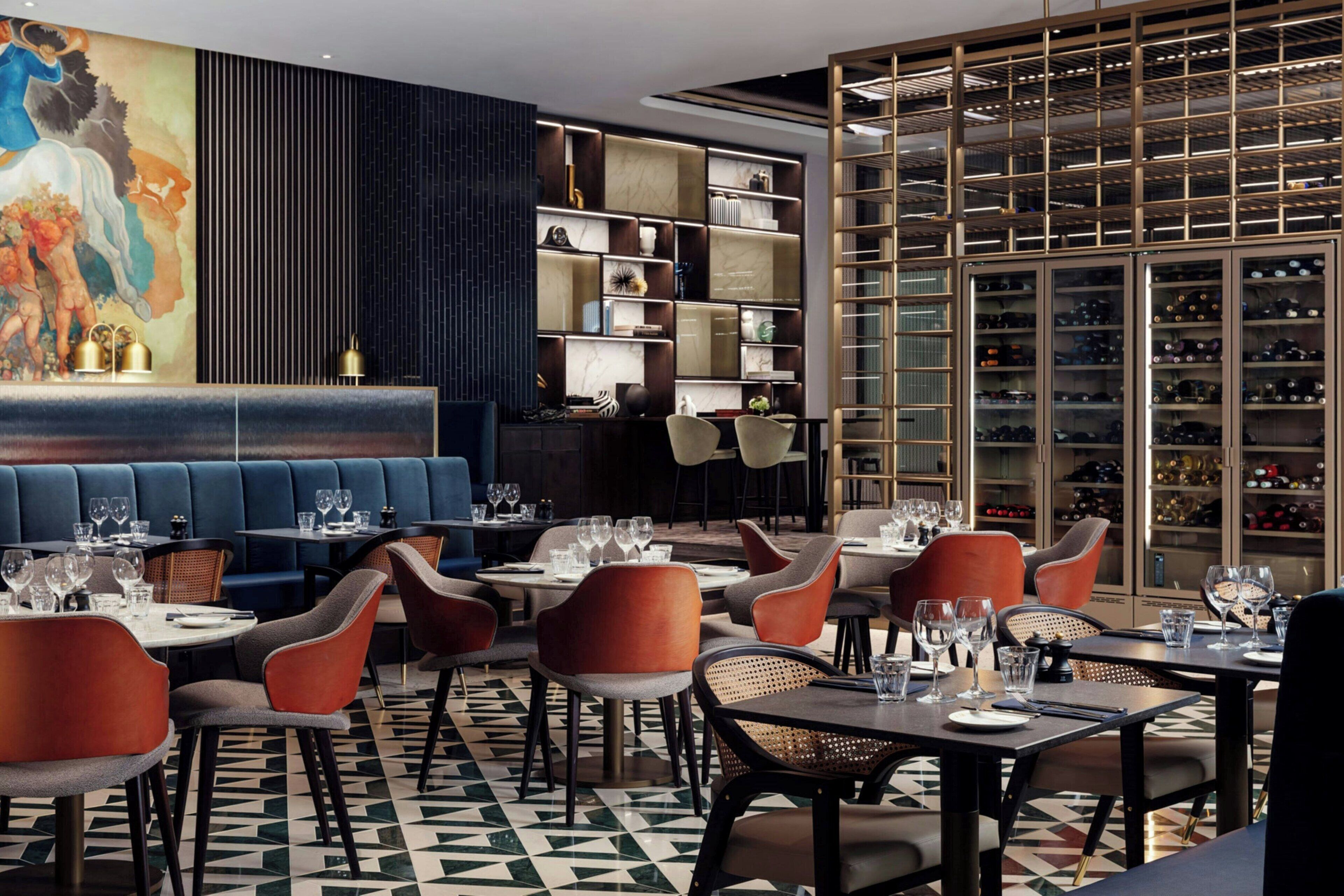 A restaurant with a geometric patterned floor, blue cushioned seating, and a wine cellar visible through a glass partition.