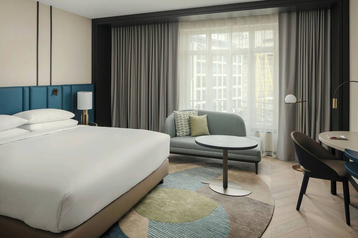 Une chambre d’hôtel avec un grand lit, un canapé, une petite table et une chaise. Il y a une fenêtre avec des rideaux et une vue sur les bâtiments à l’extérieur.