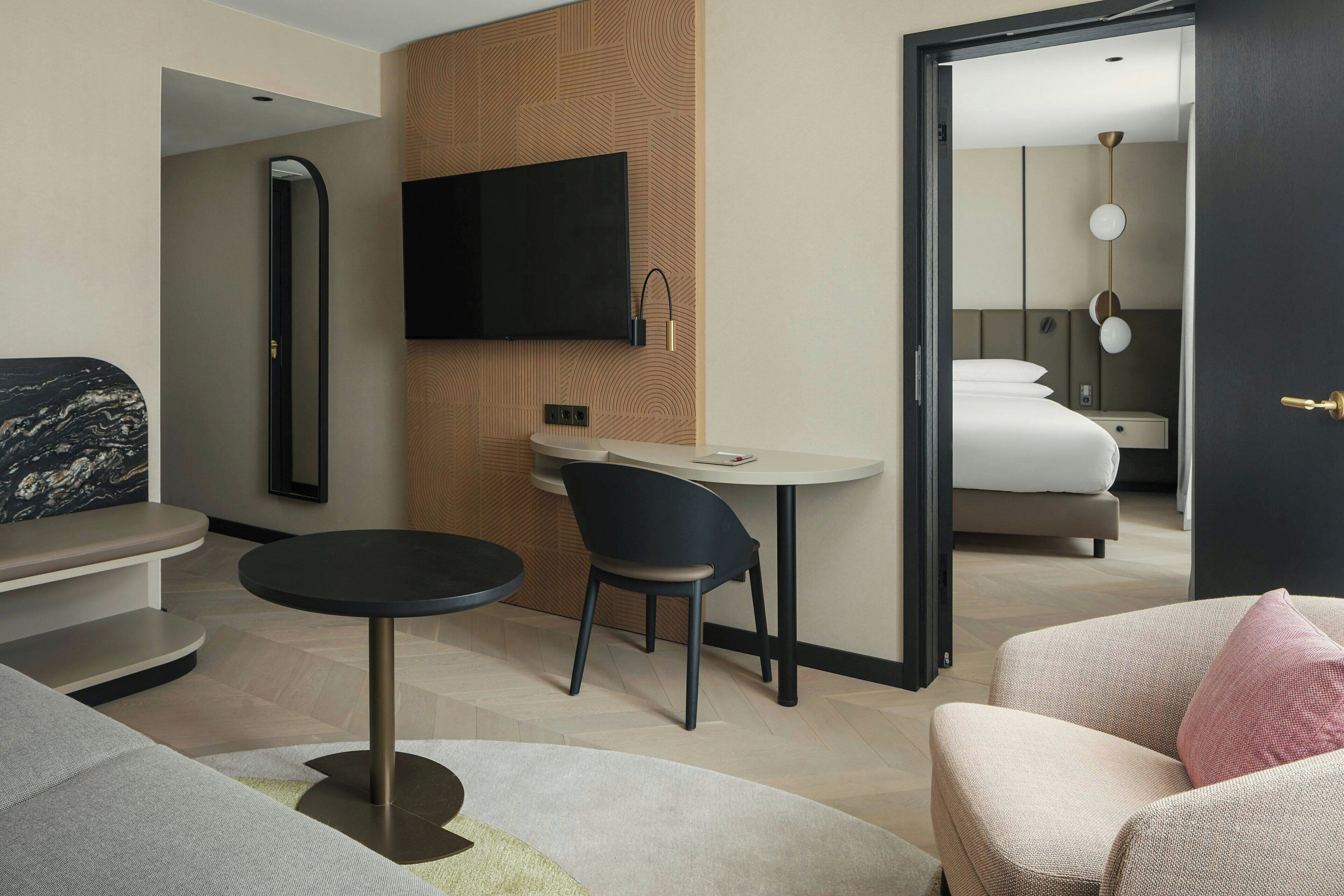 A modern hotel room with a flat-screen TV, a small round table, a dark blue chair, a sofa with a pink pillow, and a bed with white bedding.