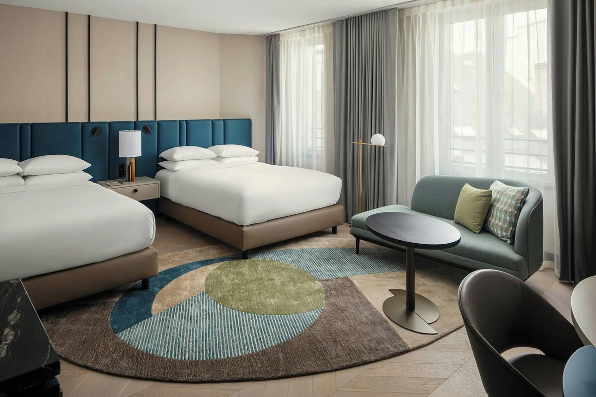 Une chambre d’hôtel moderne, dotée d’un grand lit, d’un canapé, d’une petite table et offrant une vue sur la ville grâce à de grandes fenêtres.