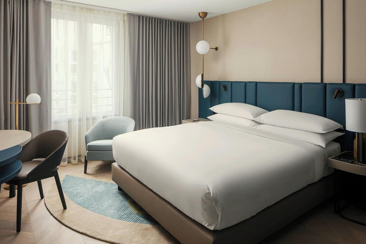 Une chambre d’hôtel avec un grand lit, un bureau avec une chaise, une lampe et une fenêtre avec des rideaux.