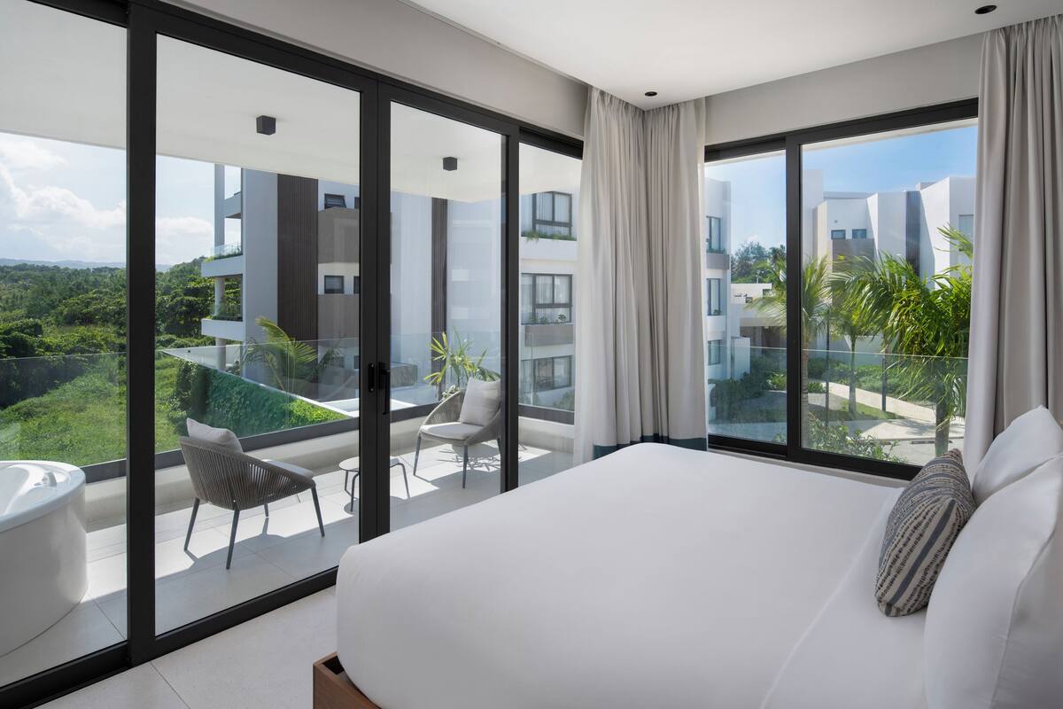 A modern hotel room with a large bed, a balcony with a chair, and a view of the city.