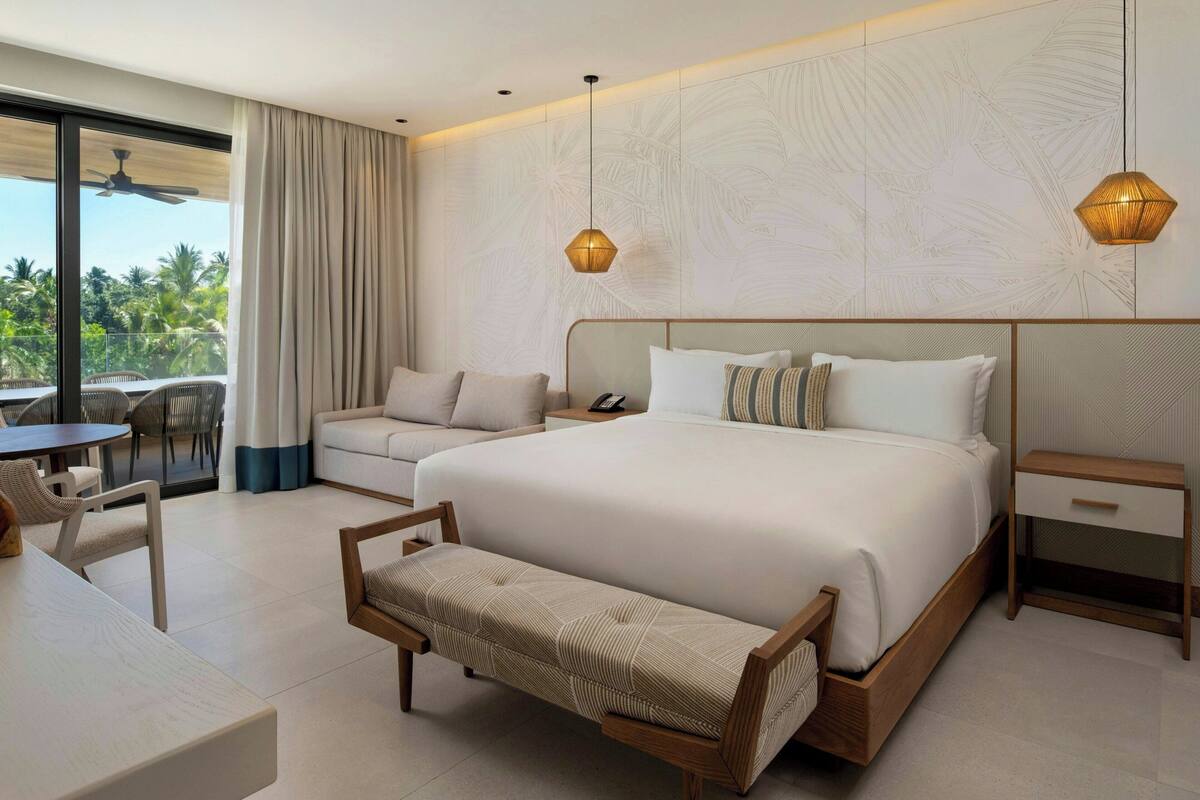 A modern hotel room with a large bed, a sofa, a dining area, and a balcony with a view of palm trees.