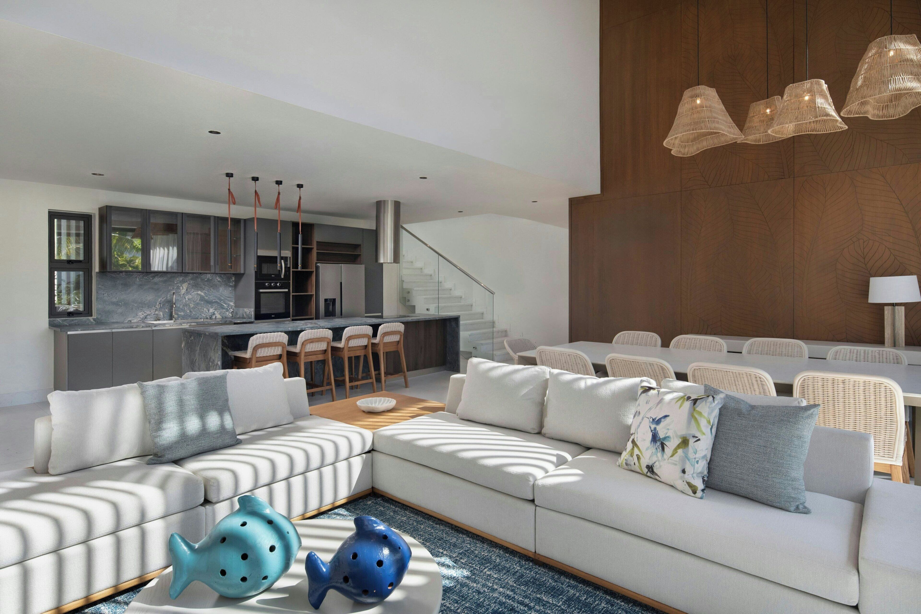 Una sala de estar moderna con un sofá modular, un comedor con sillas de mimbre blanco, y una cocina al fondo.