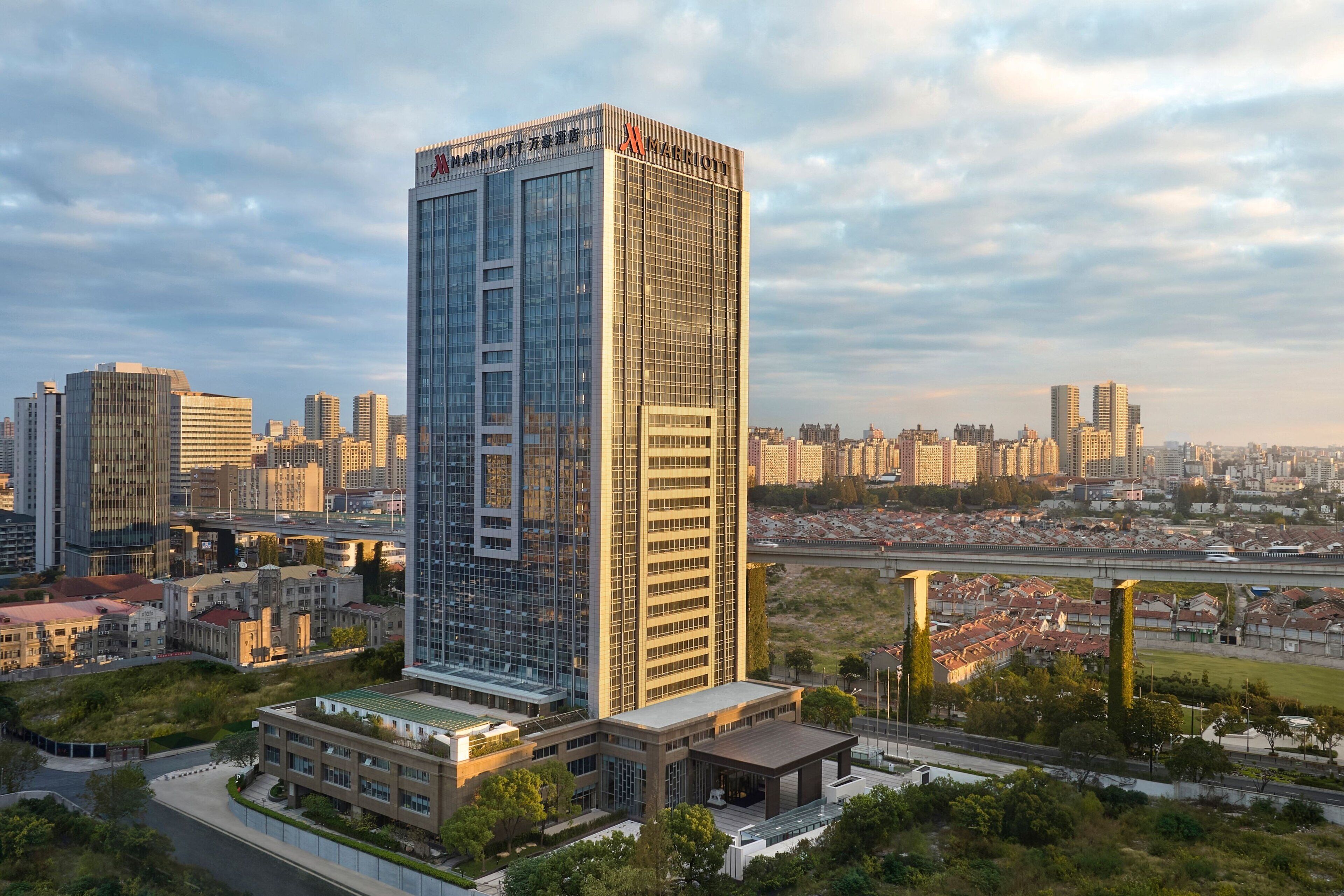 A tall, modern hotel with a glass facade, surrounded by other buildings and a green area.