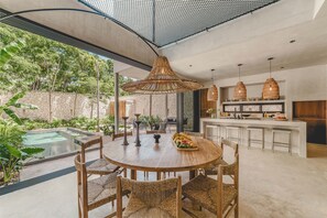 Relax in this open-plan living space featuring a central dining area with a round table and wicker chairs, adjacent to a kitchen with island and a glass door leading to a pool and lush outdoor area.
