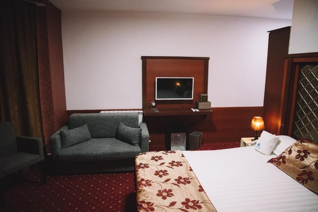 A hotel room with a bed, a sofa, a desk with a TV, and a nightstand with a lamp.