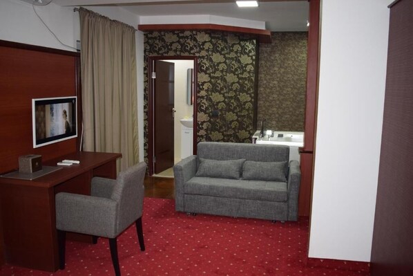 Quarto de hotel com uma escrivaninha, um sofá cinza, um tapete vermelho, uma televisão e um banheiro visível através de uma porta aberta.