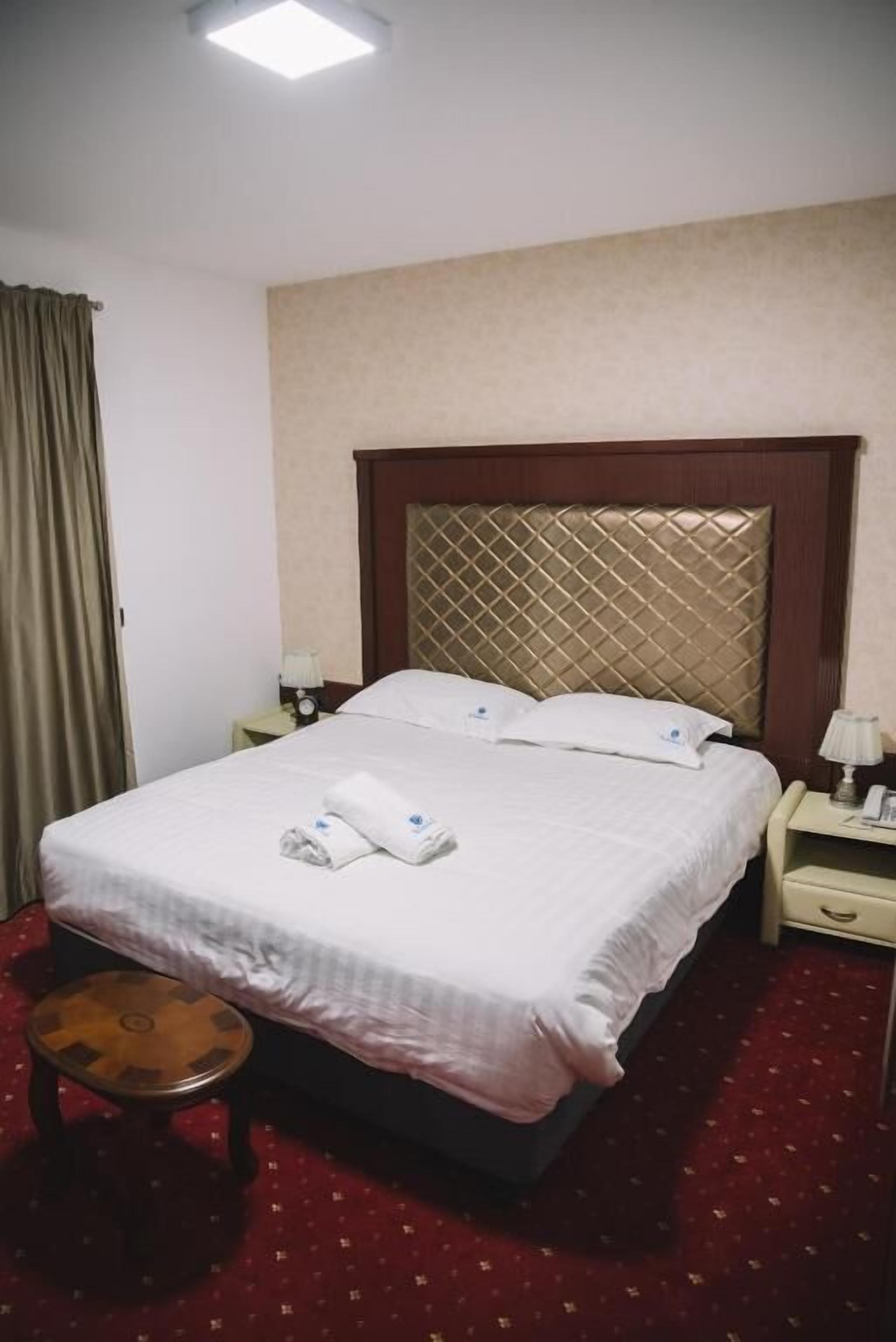 Una habitación de hotel con una cama, una mesita de noche, una lámpara, un taburete de madera pequeño y una alfombra roja.