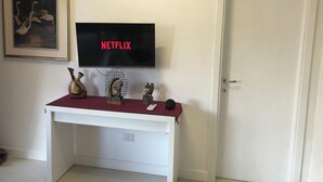 A white desk with a flat-screen TV displaying 'NETFLIX' in red letters, a framed painting, and decorative items on the desk. - Pisu Apartments (Bonarina apartment)  (Quartucciu)
