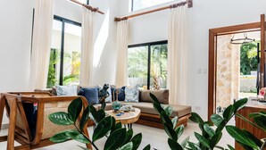 A spacious living room with a sofa, coffee table, and wicker chair. Large windows offer a view of greenery outside. - Luxury Villa for 10 Guests with Pool and Jacuzzi (Las Terrenas)