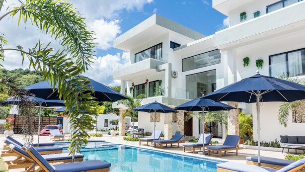 A modern pool area with lounge chairs and umbrellas, surrounded by a white building with large windows and greenery. - Luxury Villa for 10 Guests with Pool and Jacuzzi (Las Terrenas)