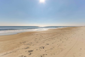 Ein weiter, sandiger Strand mit Fußabdrücken und Radspuren, der zu einem klaren Meer unter blauem Himmel führt.