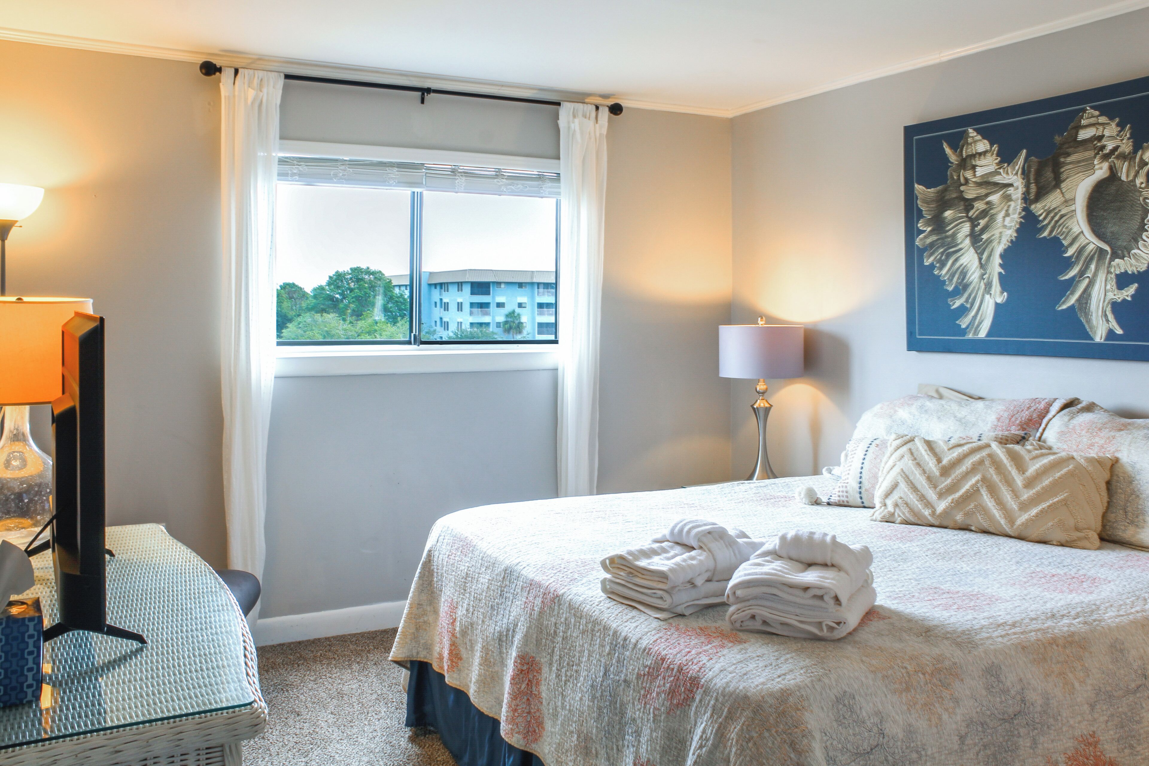 A hotel room with a bed, a window with curtains, a lamp, a wall art piece, and a folded towel on the bed.