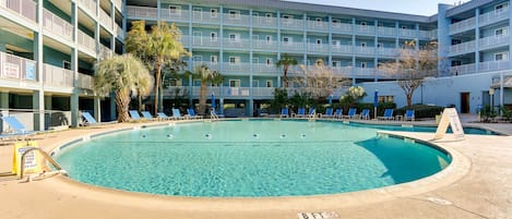 Ett hotellområde med en pool, där vattnet är kristallklart och blått, samt solstolar och en byggnad med flera våningar och balkonger.