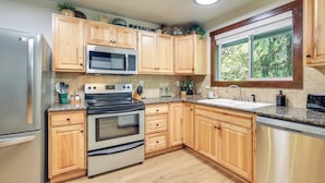 A kitchen with wooden cabinets, stainless steel appliances, a window with a view, and a countertop with various items. - Mt Hood Launchpad! Cozy Retro Rhododendron Cabin (Rhododendron)