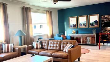 A modern living room with a brown leather sofa, a wooden coffee table, and a patterned rug. There is a ceiling fan, a blue wall with framed pictures, and a wooden cabinet.