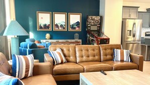 A living room with a sectional sofa, a coffee table, and a rug. There are framed pictures on the wall, a ceiling fan, and a kitchen area in the background.