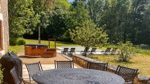 Une terrasse extérieure avec une table ronde et des chaises, un barbecue et un jacuzzi entouré de fauteuils de relaxation.