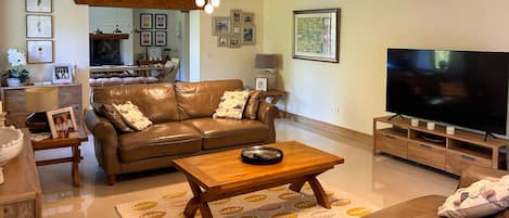 A living room with a brown leather sofa, a wooden coffee table, a television, and a dining area in the background.