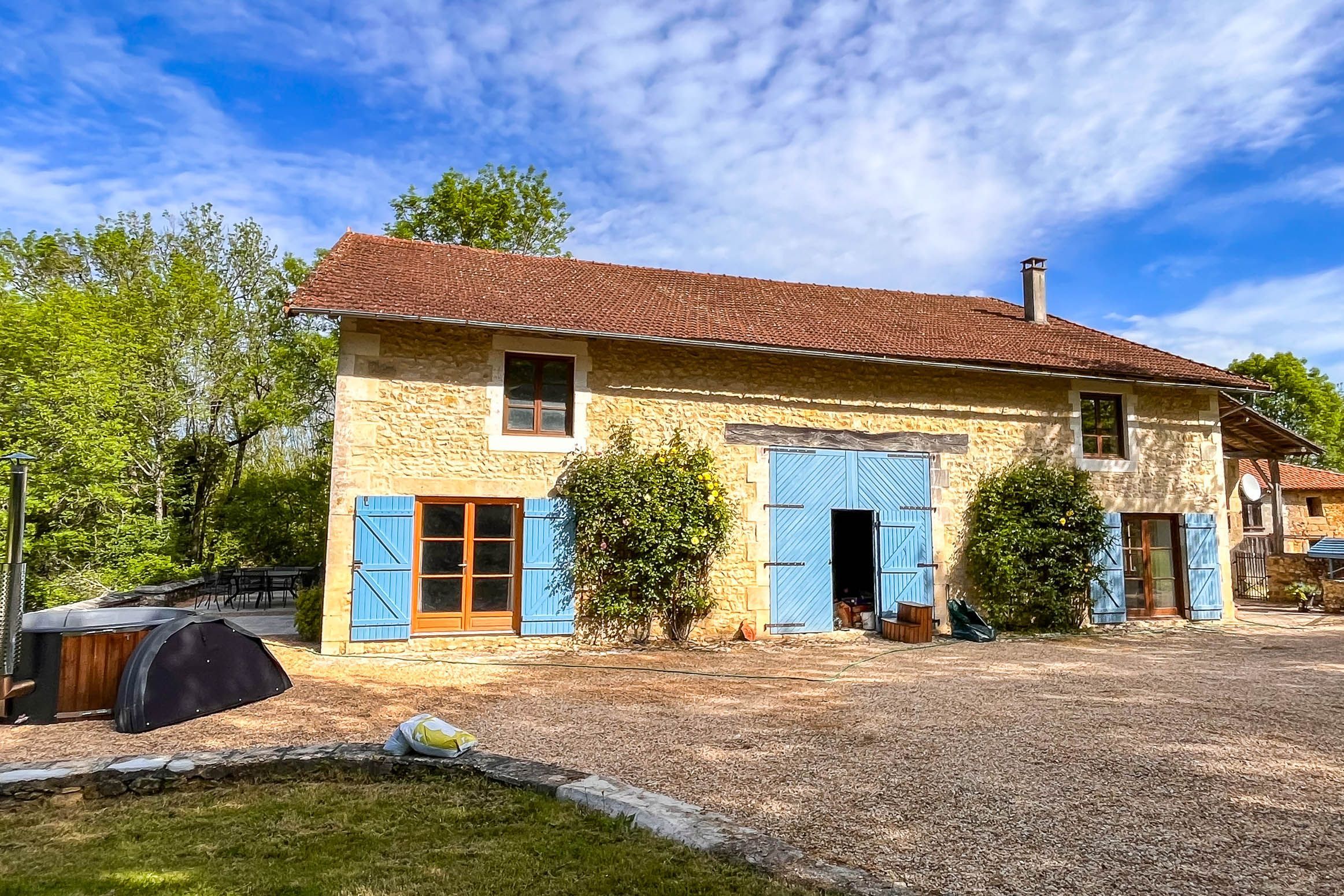 A two-story stone house with a tiled roof, a barbecue grill, and a gravel driveway.