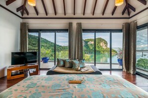 A spacious bedroom with a large bed, a television on a wooden stand, and a view of a lake and mountains through large windows. - TreeHouse Villas - Hilltop Pool Villa (Ko Yao Noi)