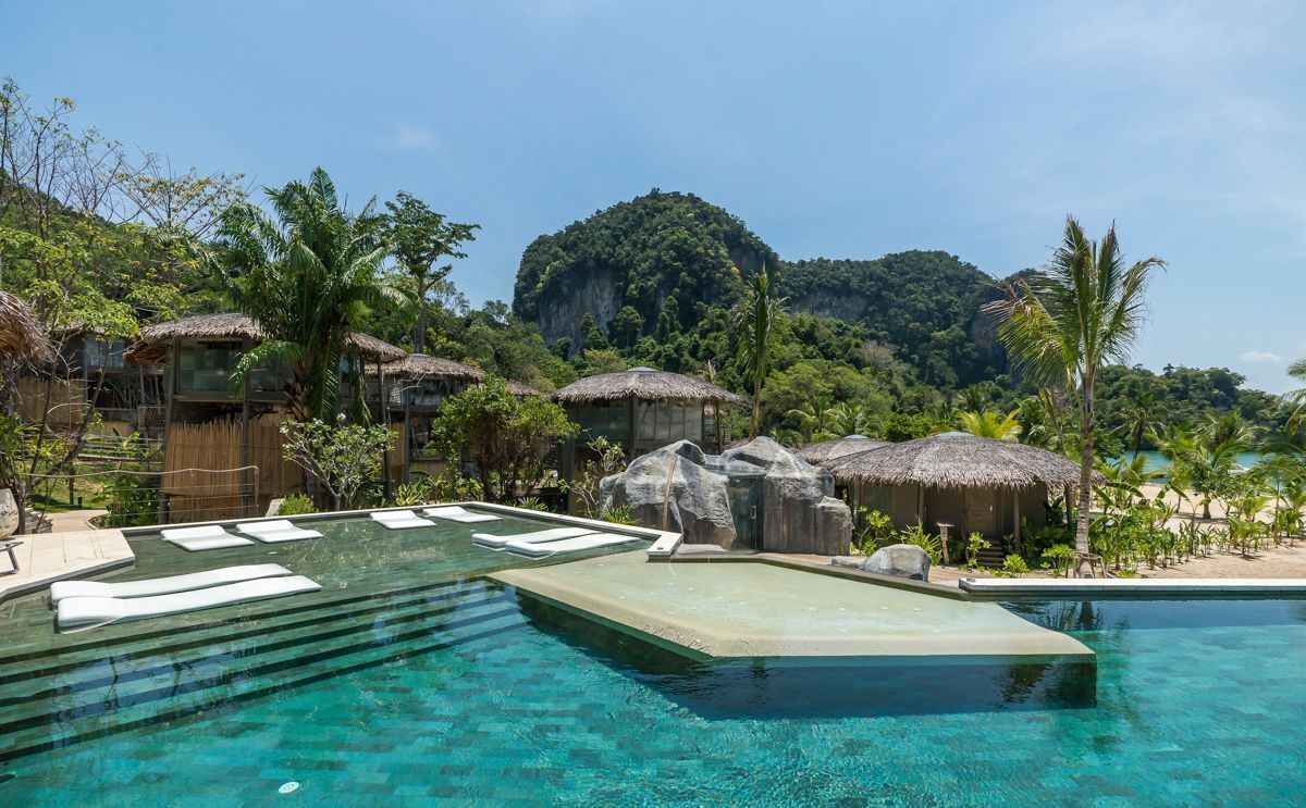 Ein klares, blaues Schwimmbecken mit Liegestühlen, umgeben von Hütten mit Strohdächern und Palmen, mit einem Berg im Hintergrund.