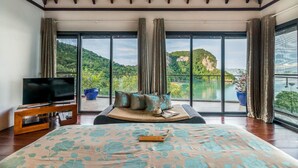 A spacious bedroom with a large bed, a television on a wooden stand, and a view of a lake and mountains through large windows. - TreeHouse Villas - Hilltop Pool Villa (Ko Yao Noi)