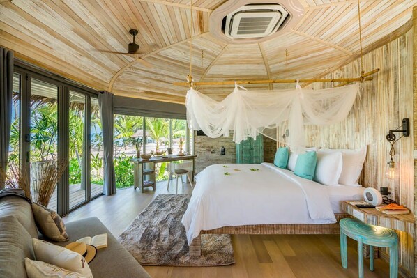 A spacious bedroom with a large bed, a sitting area with a table and chairs, and a view of greenery through large windows. - TreeHouse Villas - Beachfront Pool Villa (Ko Yao Noi)