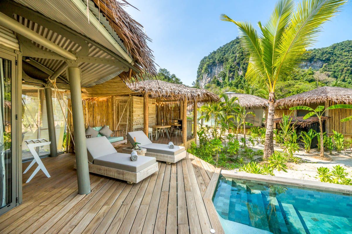 A wooden deck with lounge chairs and a pool, surrounded by thatched-roof huts and palm trees.