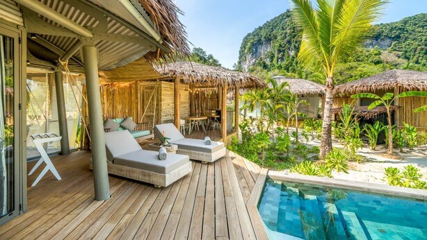 Une terrasse en bois avec des chaises longues et une piscine, entourée de huttes à toits de chaume et de palmiers.