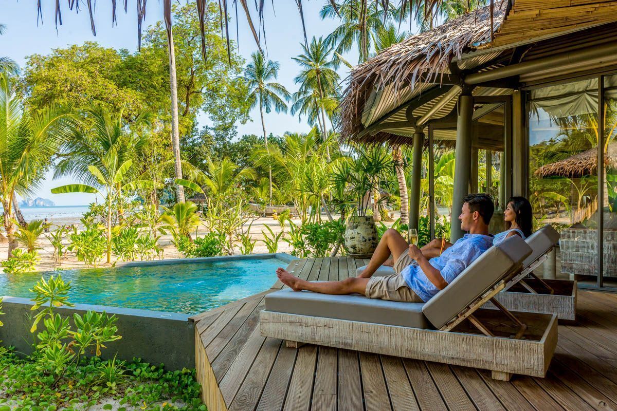 Un terrazzo in legno con poltrone relax che si affaccia su una piscina, circondato da vegetazione tropicale e palme.