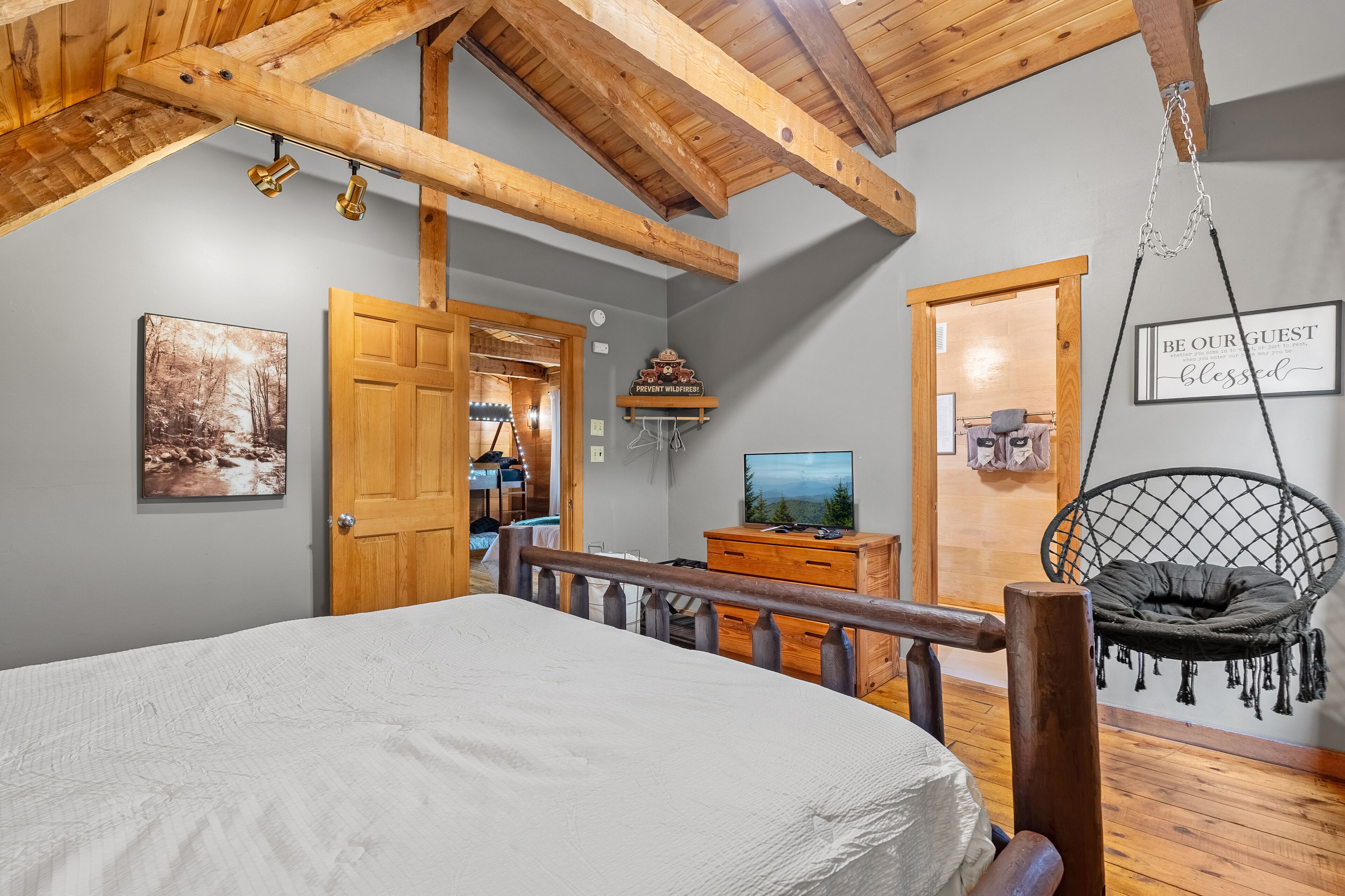 A bedroom with a bed, wooden ceiling beams, a hanging chair, a television, and a door leading to another room.