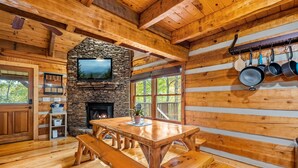 Interior rústico de cabaña con chimenea de piedra, mesa de comedor de madera, bancos y pared de piedra con un televisor.