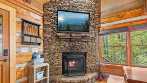 Una chimenea de piedra con un televisor encima, paredes de madera y vistas a árboles a través de una ventana.