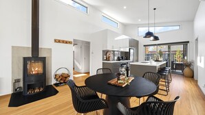 A modern kitchen with a central island, a dining area with a round table and chairs, a built-in fireplace, and large windows providing natural light.