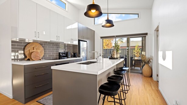 A modern kitchen with a large island, dark grey cabinets, and a black countertop. There are three black bar stools, a wooden cutting board, and a coffee machine.