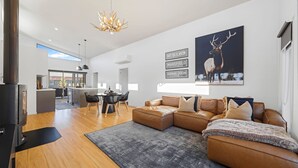 A modern living room with a sectional sofa, a dining area with a table and chairs, a kitchen in the background, and a large deer painting on the wall.
