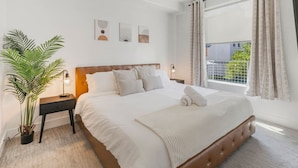 Un dormitorio con cama, ventilador de techo, mesita de noche con lámpara, planta en maceta y ventana con cortinas.
