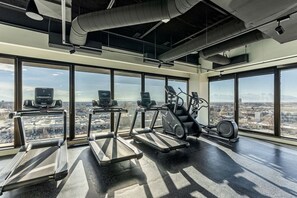 A gym with treadmills, elliptical machines, and a stationary bike, large windows offering a city view.