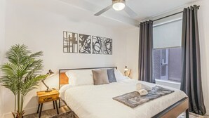 A bedroom with a bed, a ceiling fan, a window with curtains, a bedside table with a lamp, and a plant.